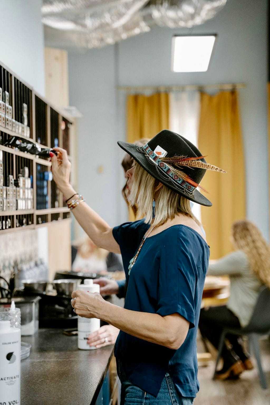 Woman in hat reaching for essential oils in a cozy shop setting.