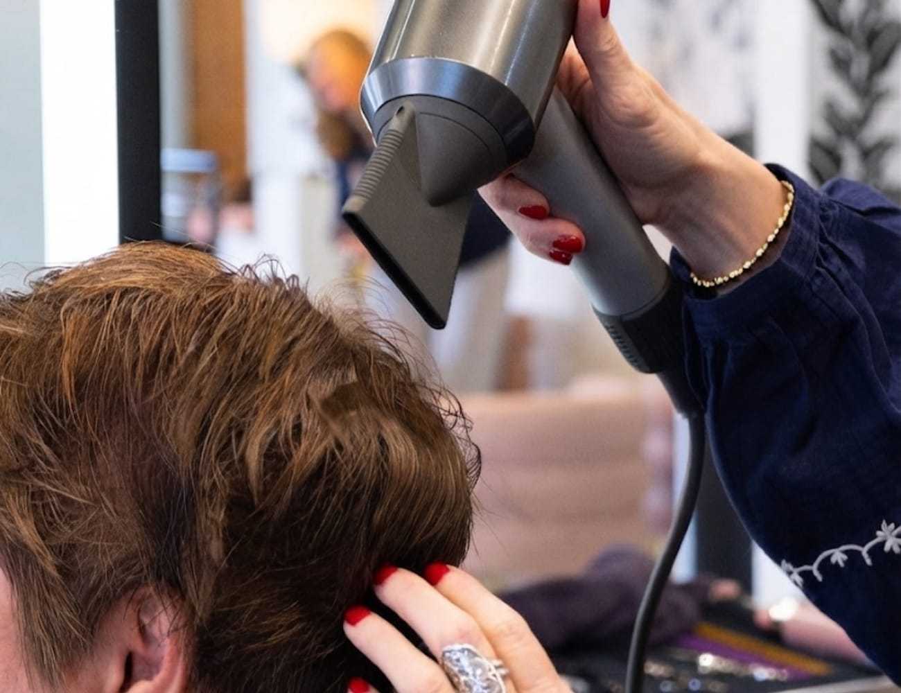 Hair being blow-dried in a salon, held by a person with red nail polish.